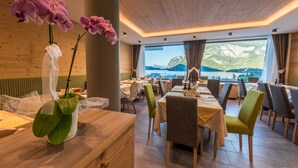 A restaurant with wooden walls and large windows overlooking a mountainous landscape. Tables are set with chairs, and there are orchids on the counter. - Feeling Hotel Fontanella (Molveno)