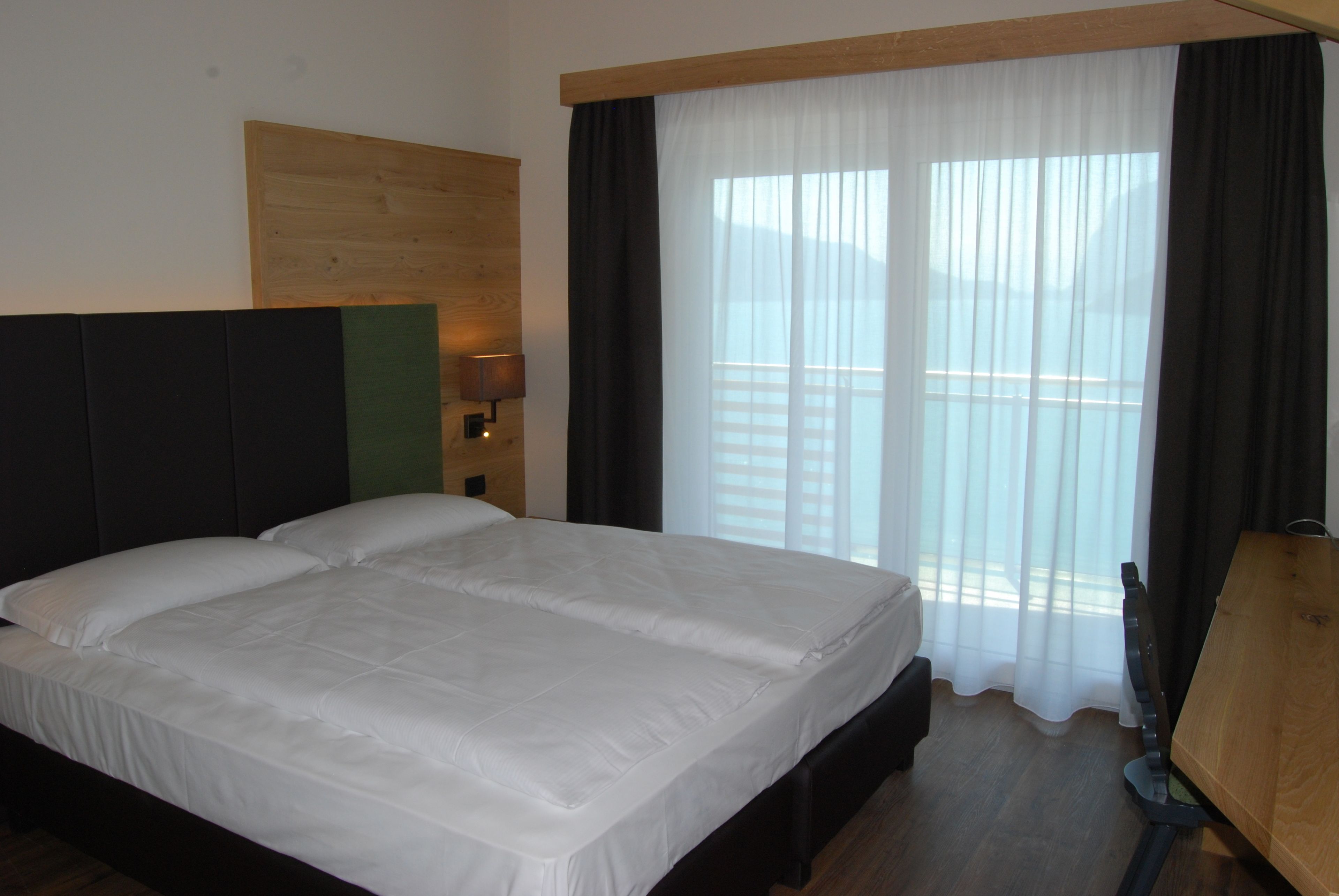 Una cama doble con ropa blanca, cabecera de madera, mesita de noche con lámpara y vistas al exterior a través de cortinas traslúcidas.