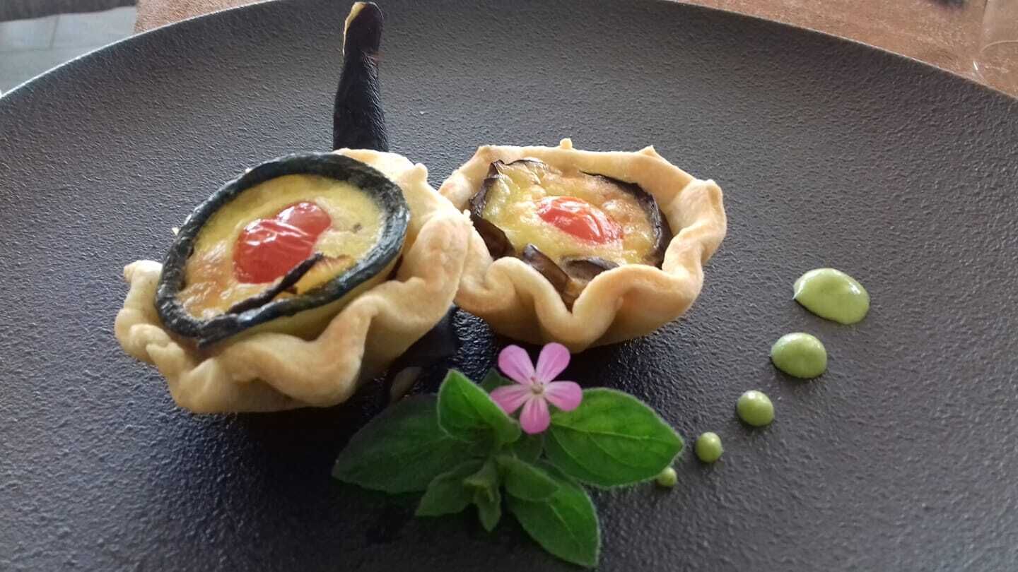 Two tartlets with a cherry on top, placed on a dark plate with green leaves and small green spheres.