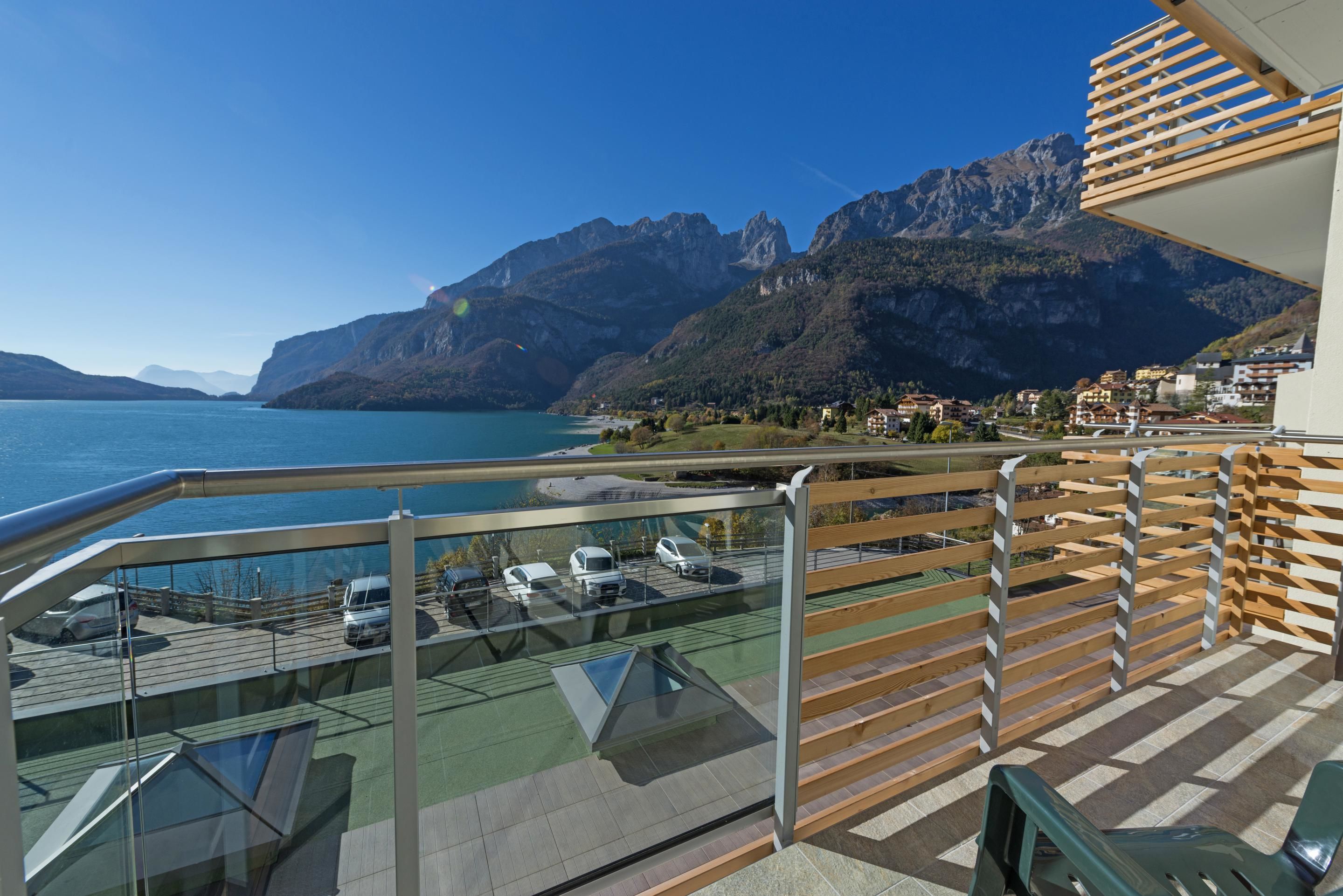 A balcony with a view of a lake and mountains, a parking area with cars, and a wooden fence.