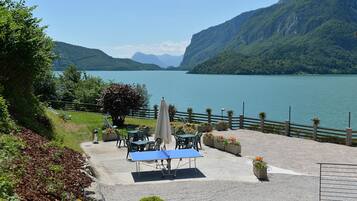 Un patio junto al lago con mobiliario exterior y una mesa, rodeado de vegetación y con vista a un lago y montañas al fondo.