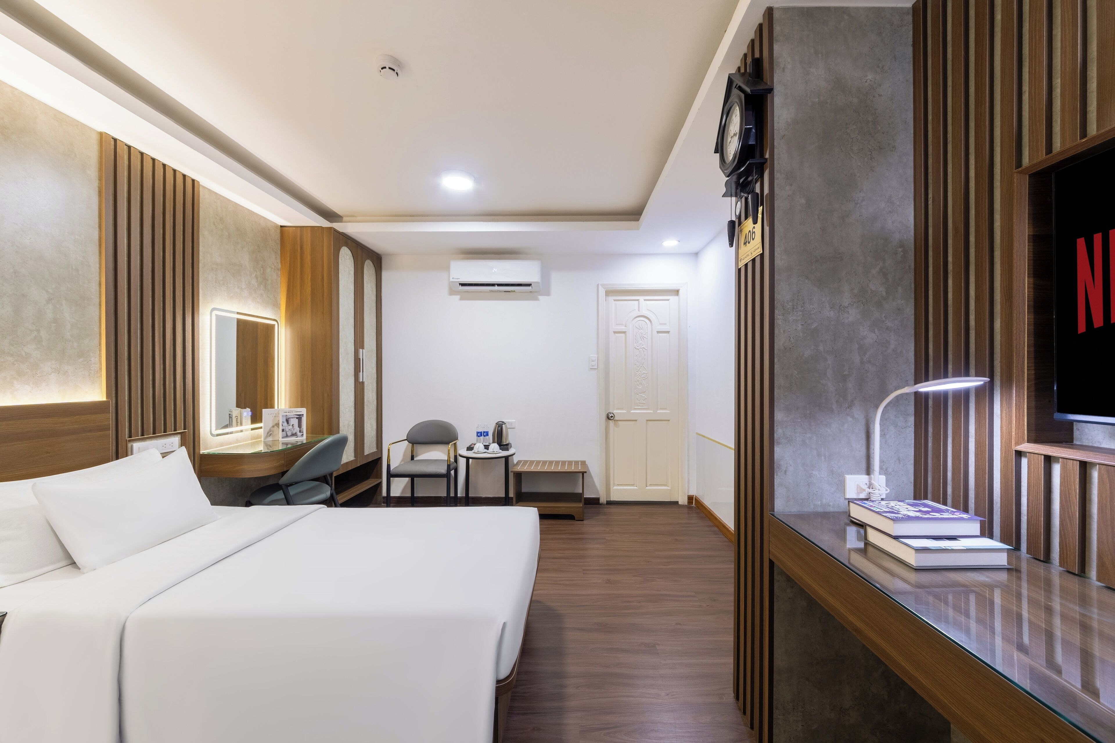 Habitación de hotel con una cama, un escritorio con lámpara, una silla, una mesita con tetera y un televisor.