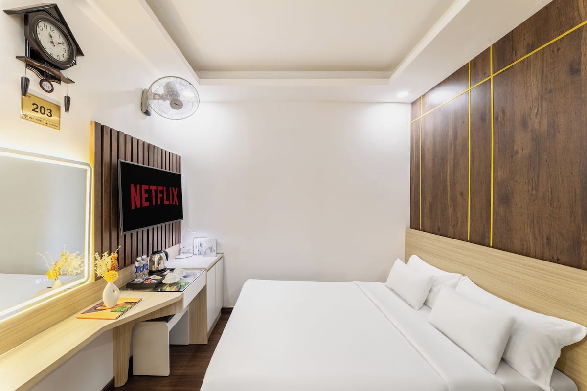 Ein Hotelzimmer mit einem Bett, einem Schreibtisch mit Computer, einer Uhr, einem Fernseher, der 'NETFLIX' anzeigt, und einem Wandventilator.