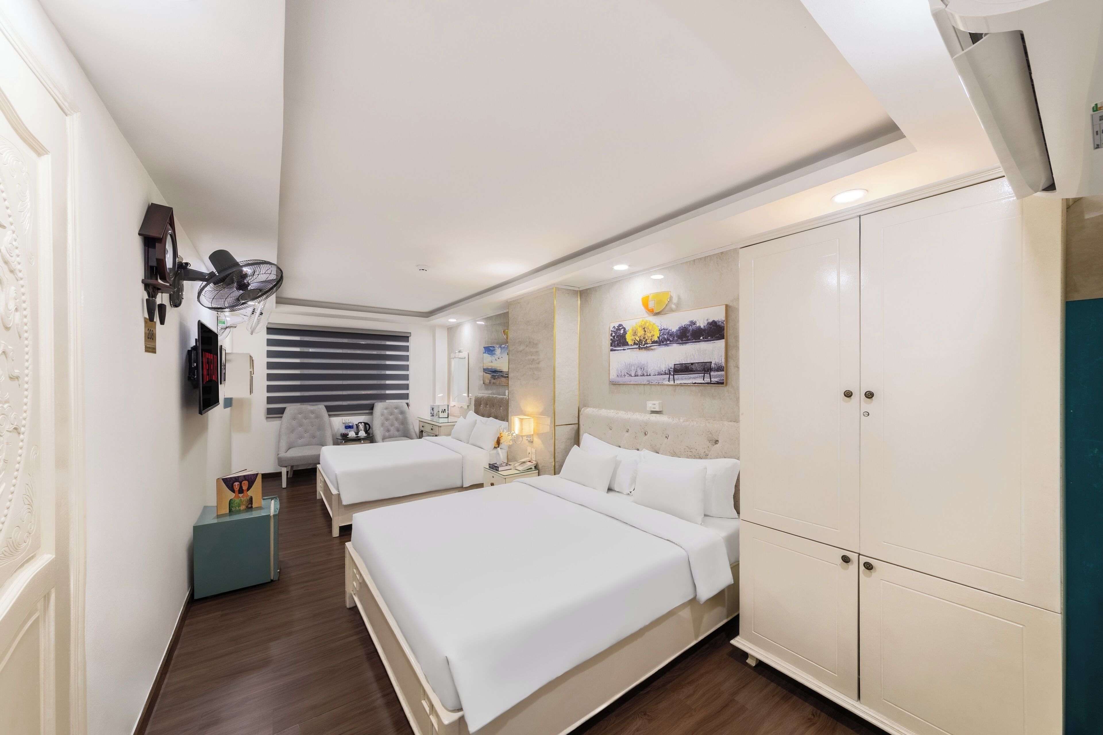 Habitación de hotel con dos camas, un armario, un escritorio, una silla, un ventilador y un cuadro en la pared.