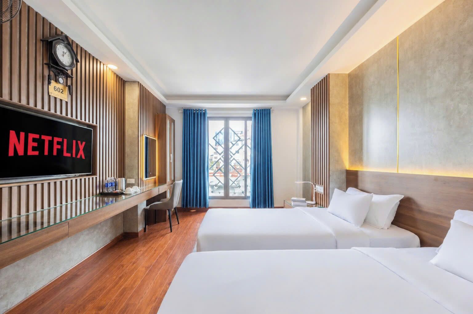 A hotel room with a large TV screen displaying 'NETFLIX,' a wooden desk, two beds, a chair, and a window with blue curtains.