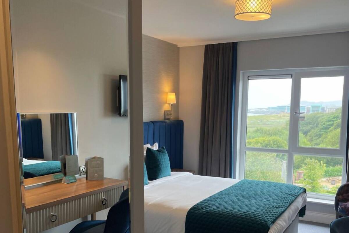 A hotel room with a bed, a desk, a chair, a TV, a window with a view, and a lamp.