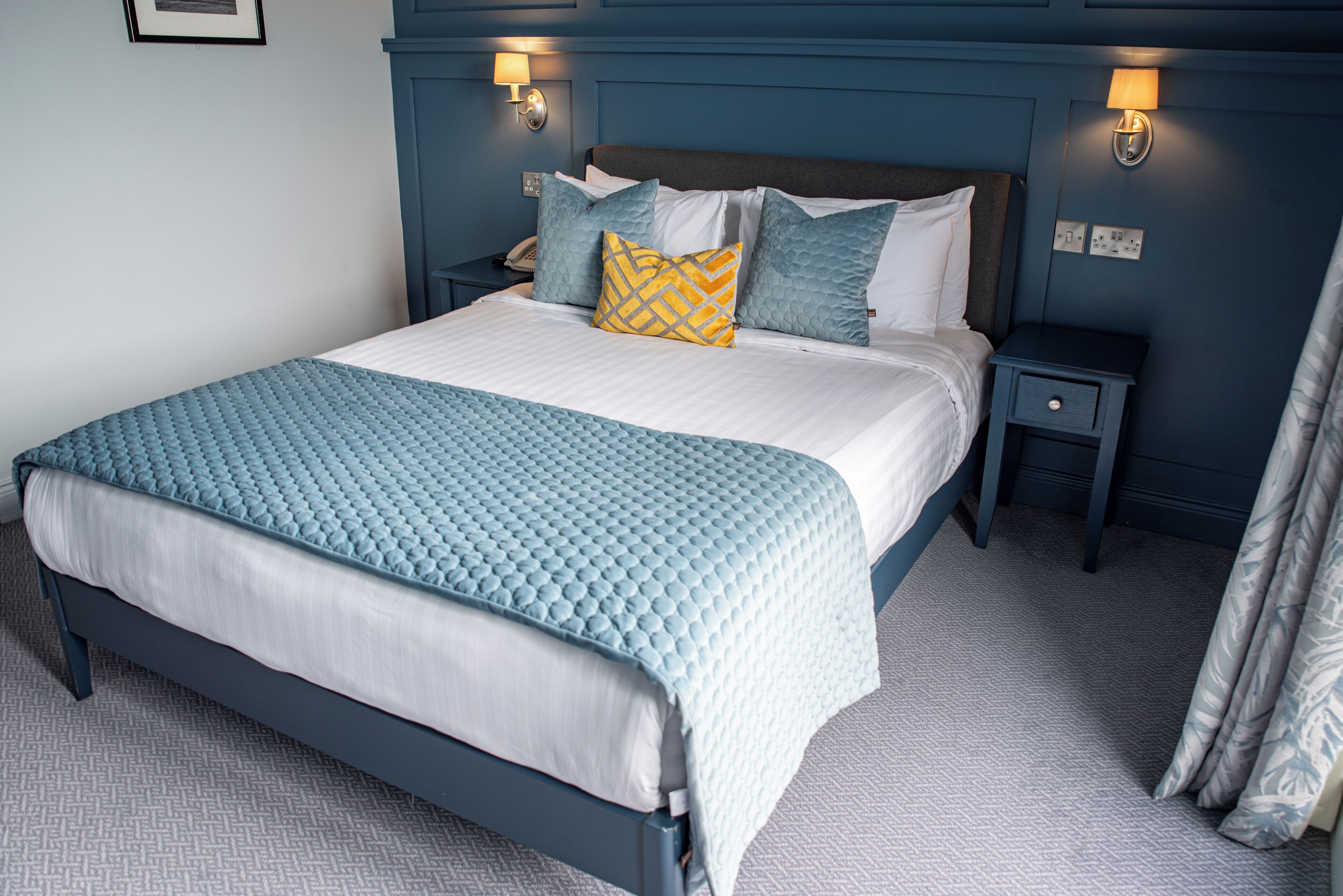 A neatly made bed with a blue quilt, a white comforter, and several pillows in shades of blue and yellow. There are two bedside tables with lamps, and the room has a blue wall and a patterned carpet.