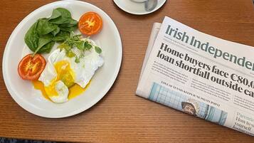 A breakfast table with a plate of eggs, tomatoes, and greens, a cup of tea, a glass of orange juice, and a newspaper.