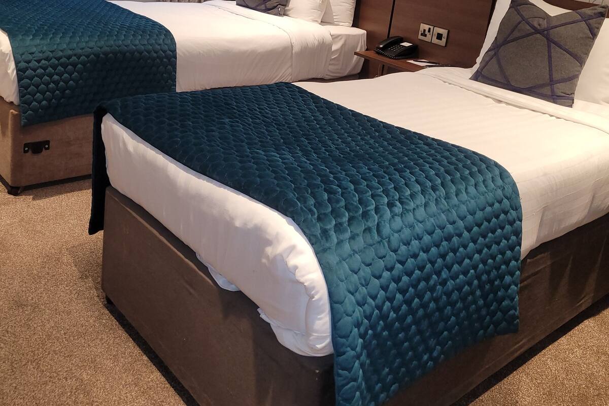 A hotel room with two beds, each with a teal quilted bedspread and white linens. There is a window with sheer curtains and a patterned wall.