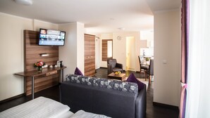 A modern hotel room with a sofa, a TV mounted on the wall, a small table with a fruit bowl, and a desk with a vase of flowers. - BATU Apart Hotel (Munich)