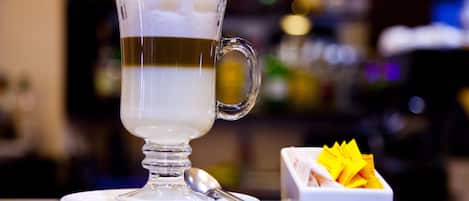 A clear glass cup of a layered beverage on a saucer with a spoon beside it, and a small white dish with yellow items in the background.