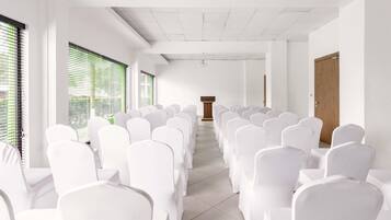 Un salón de conferencias con hileras de sillas blancas, un atril y ventanas grandes con persianas.