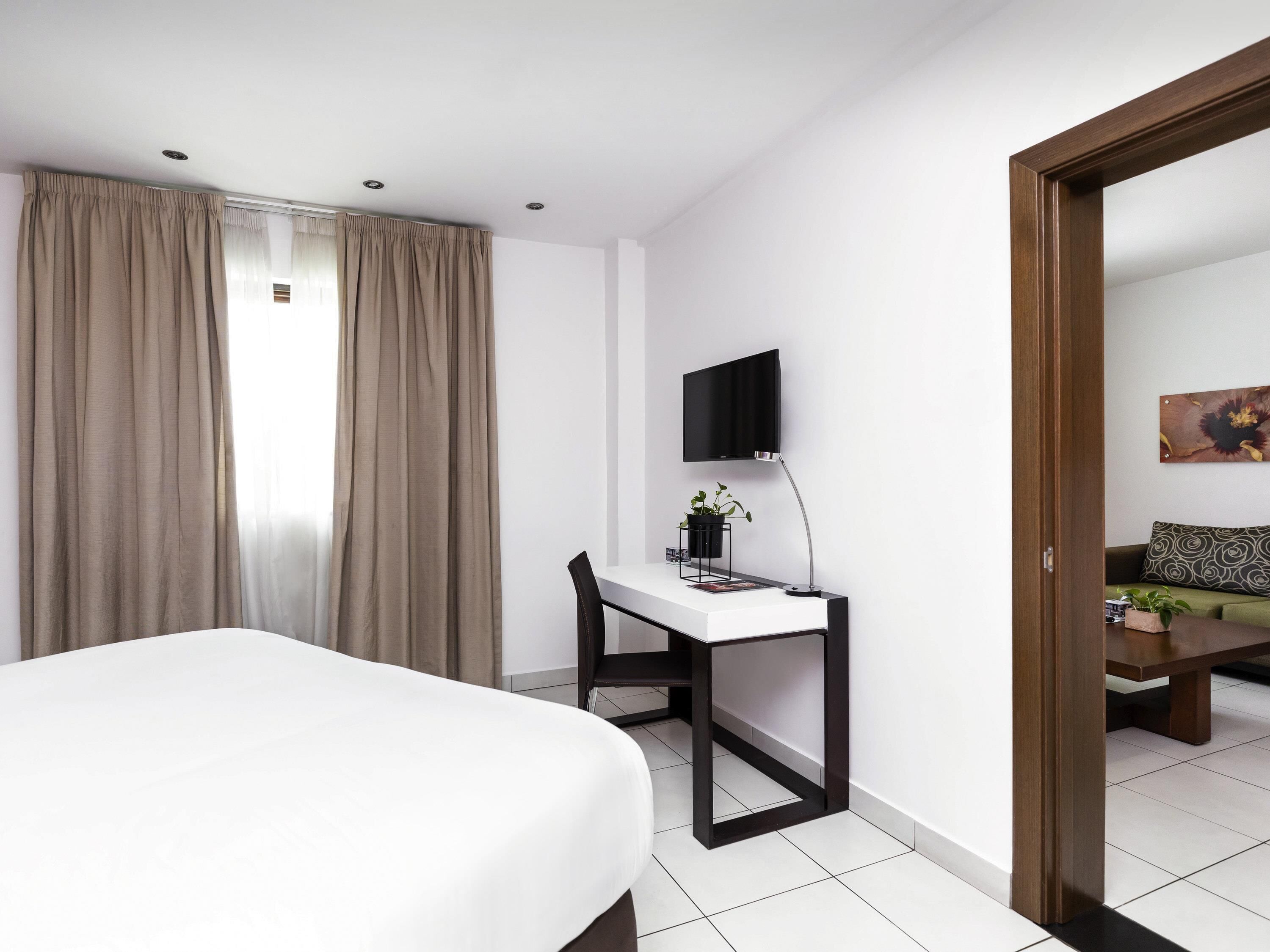 A hotel room with a bed, a desk with a chair, a TV mounted on the wall, a window with curtains, and a door leading to another room.