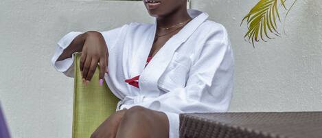A person seated on a chair outdoors, wearing a white robe, with greenery in the background.