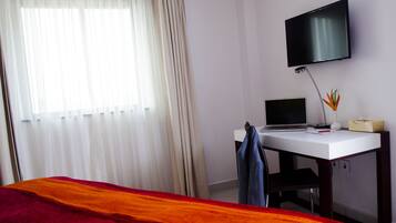 A hotel room with a bed, a desk with a laptop, a TV on the wall, and a window with curtains.