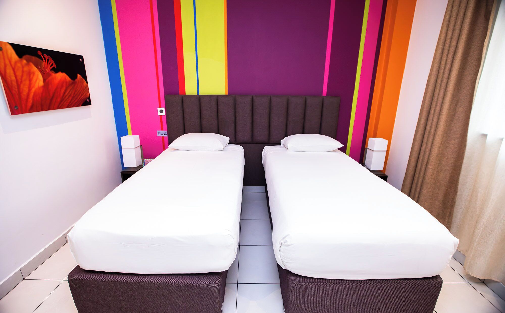 Une chambre d’hôtel avec deux lits simples, une grande fenêtre et un mur décoré de motifs colorés.