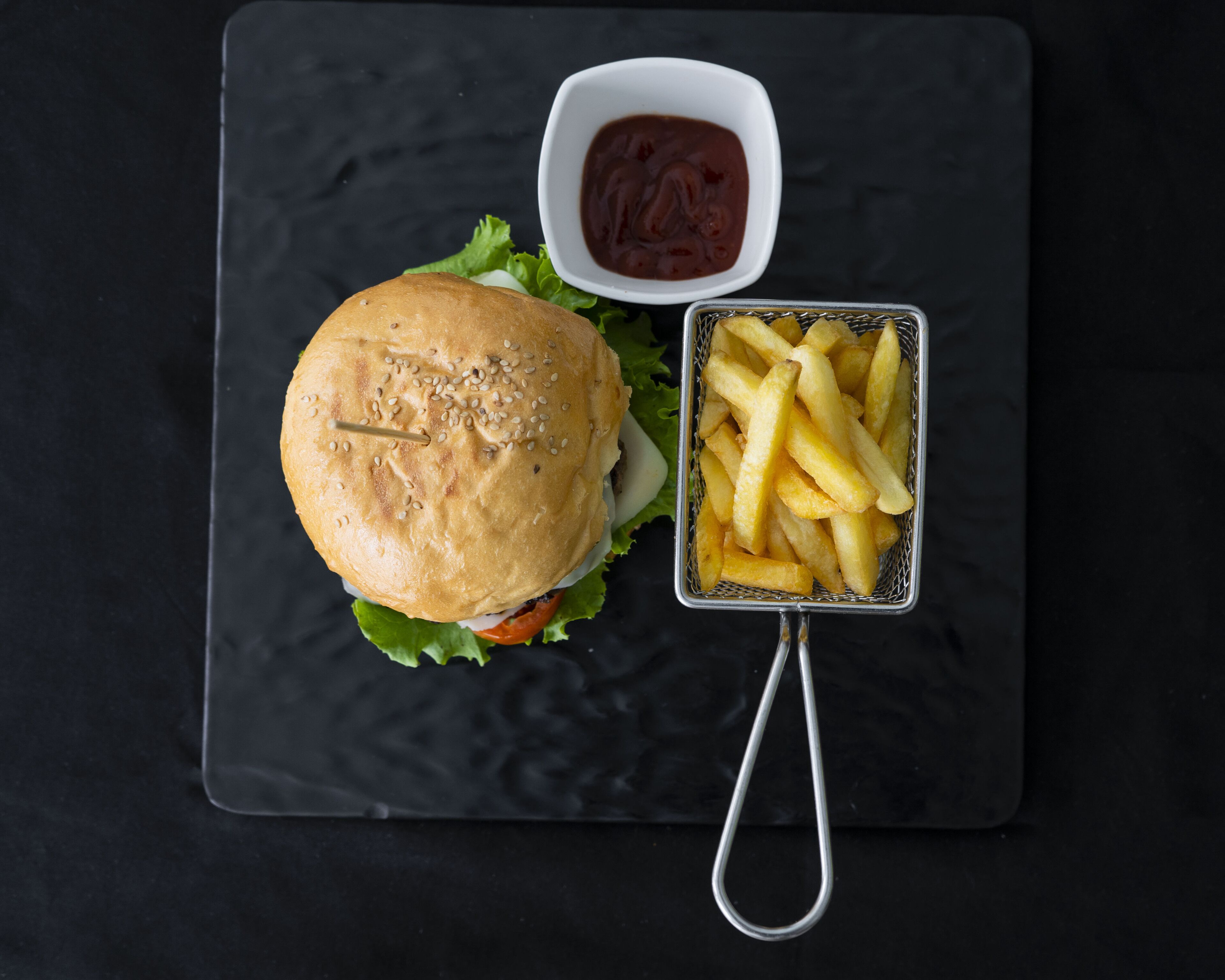 A burger with lettuce, a small bowl of ketchup, and a basket of French fries on a black slate board.