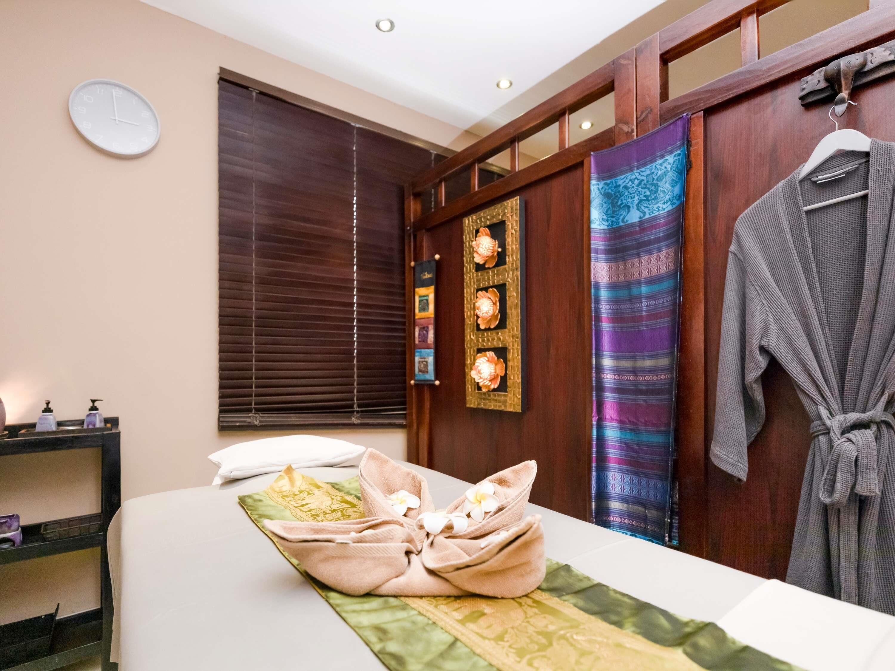 A hotel room with a bed, a robe, a towel, a clock, and a window with blinds.