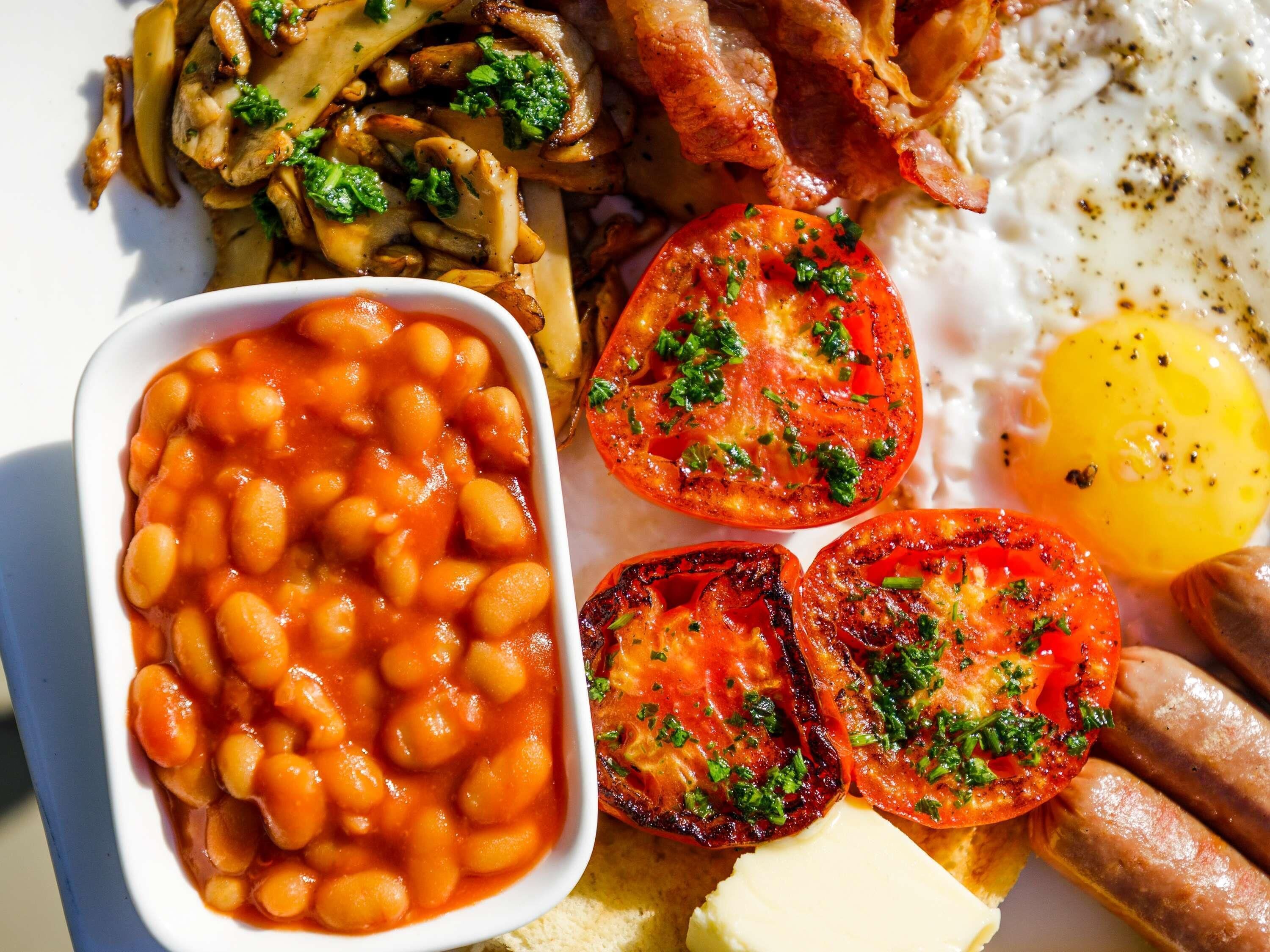 Un plato de desayuno con frijoles, tomates, huevos, tocino, salchichas, champiñones y tostadas.