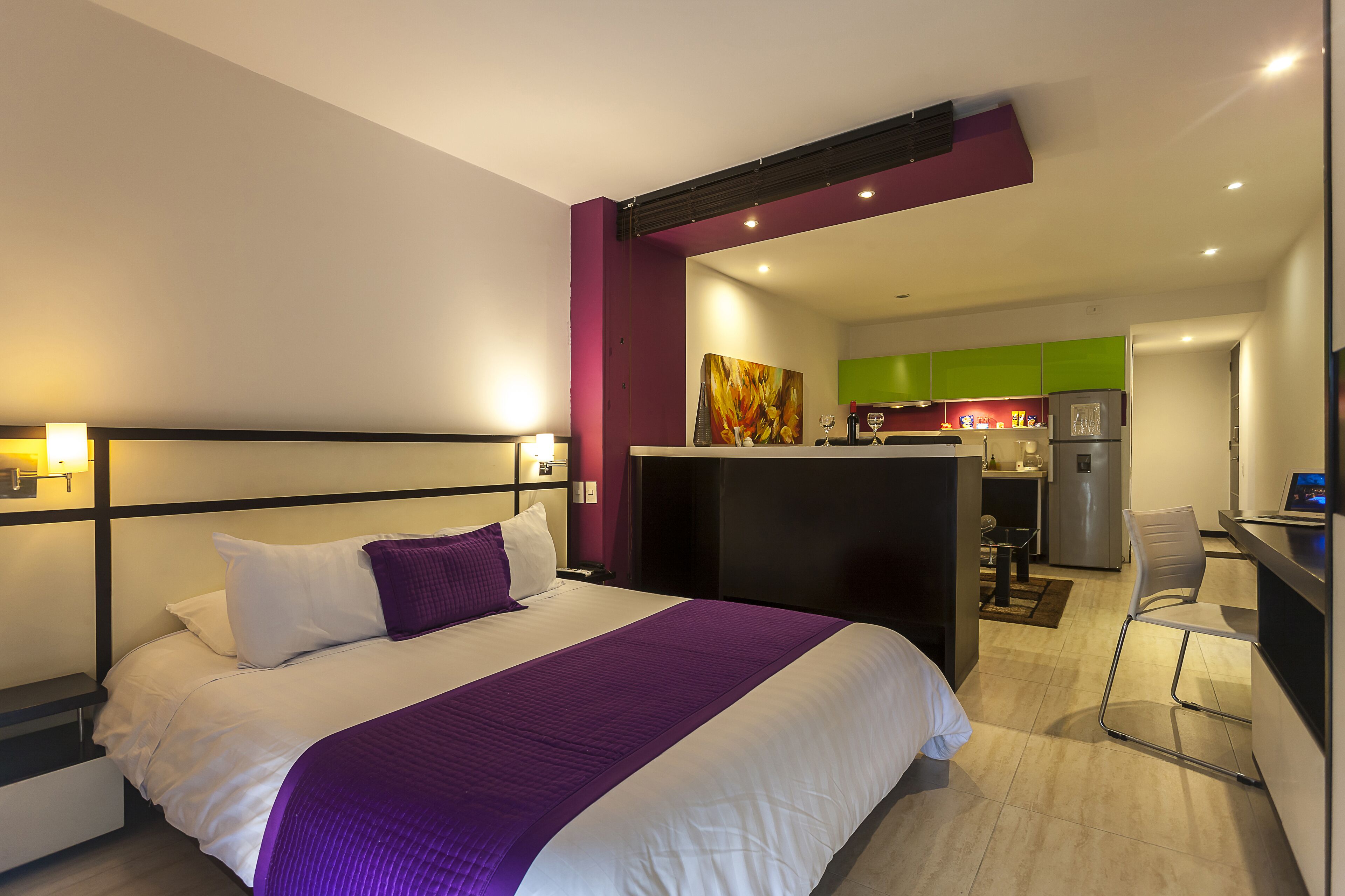 A modern hotel room with a large bed, a desk, and a kitchenette in the background.