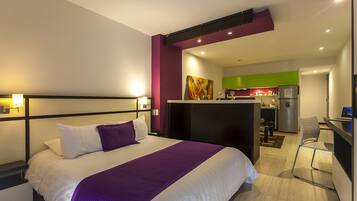A modern hotel room with a large bed, a desk, and a kitchenette in the background.