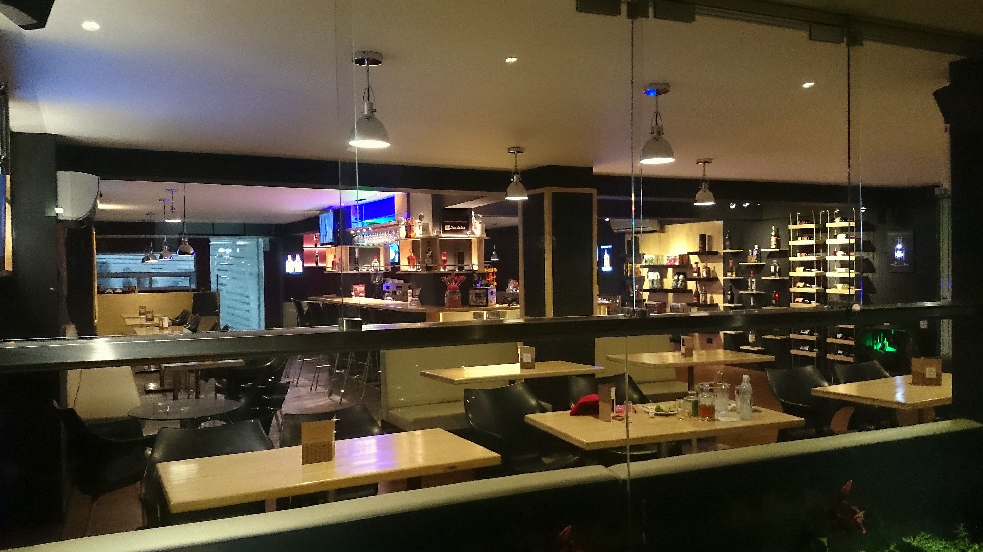 An indoor restaurant with multiple tables and chairs, a bar area with shelves of bottles, and hanging pendant lights.