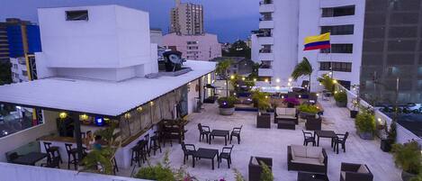 Un espacio al aire libre con patio, mobiliario, piscina, edificios circundantes y una bandera.