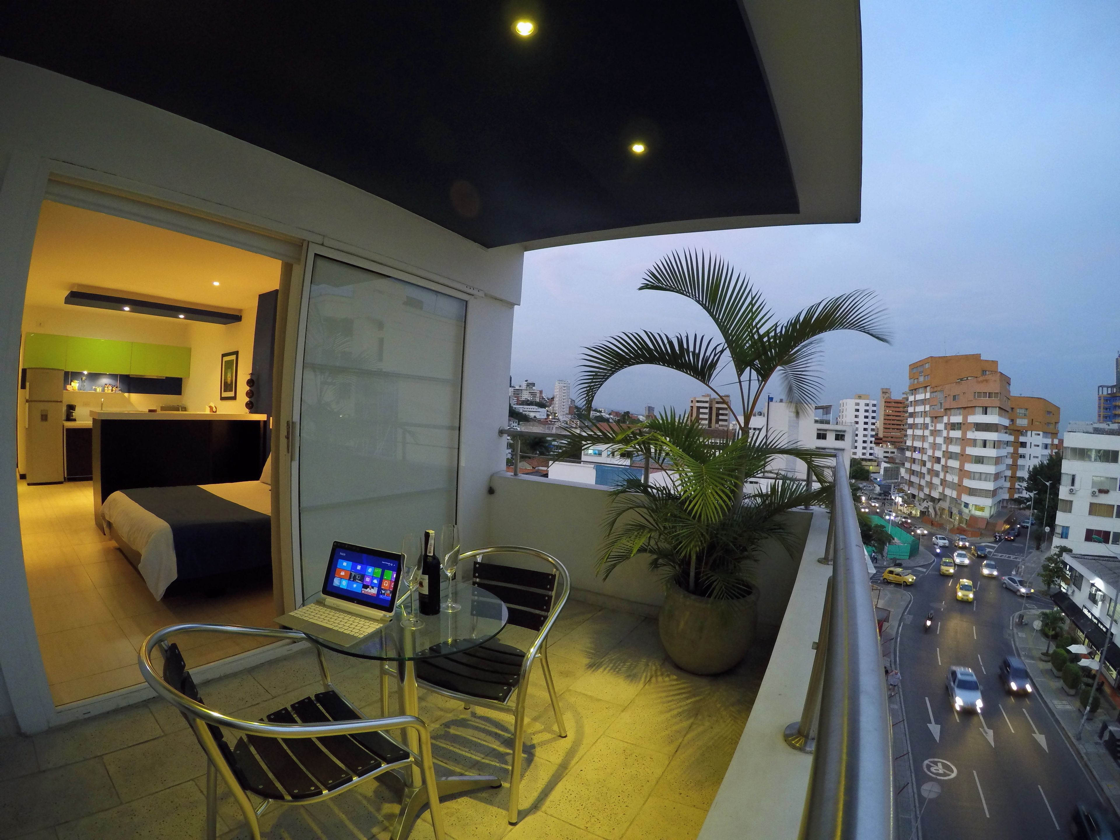 A balcony with a laptop, a glass table, and chairs, overlooking a cityscape with buildings and a road.