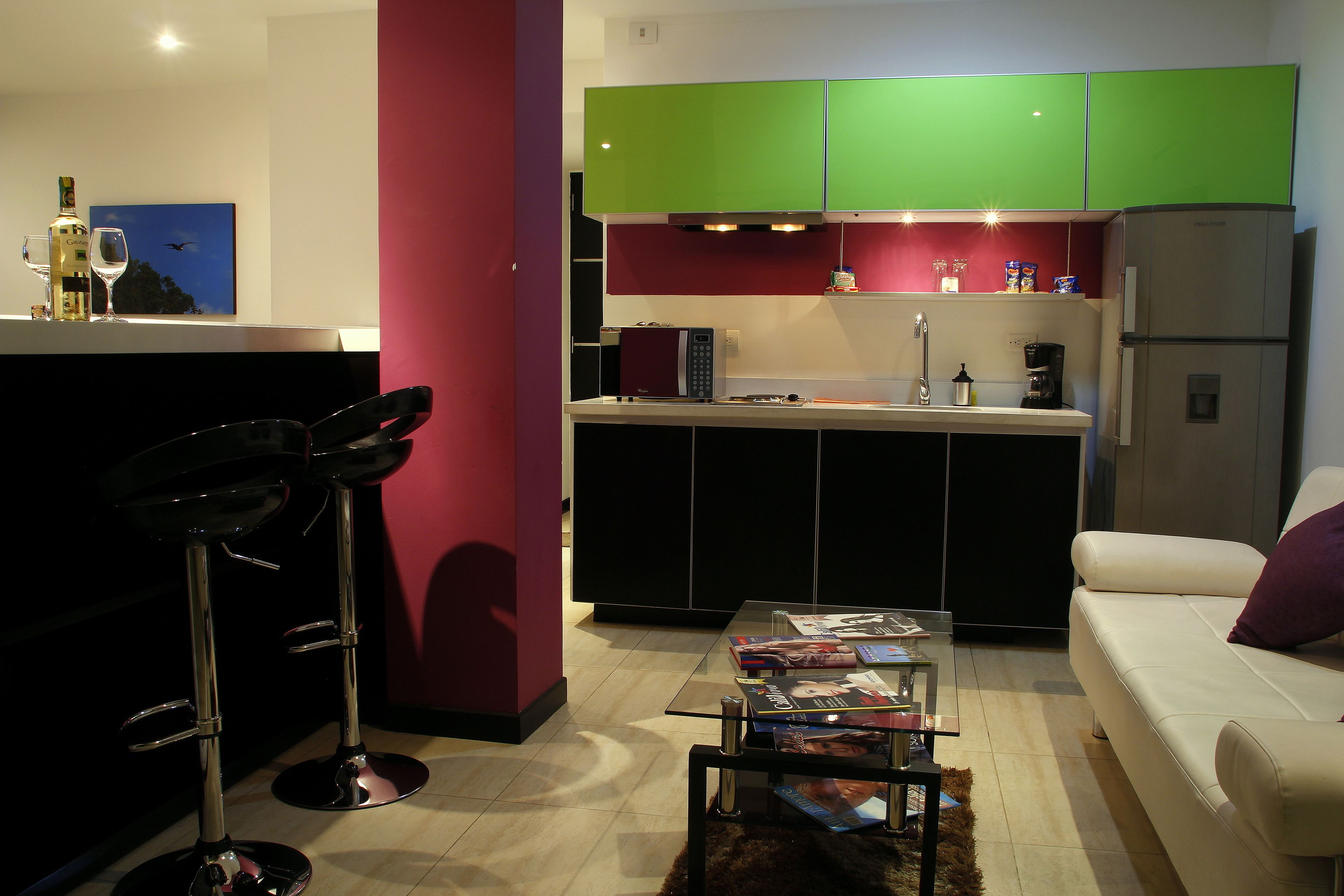 A modern kitchen with green cabinets, a white countertop, and a built-in microwave. There is a white sofa with a purple pillow and a glass coffee table with magazines.