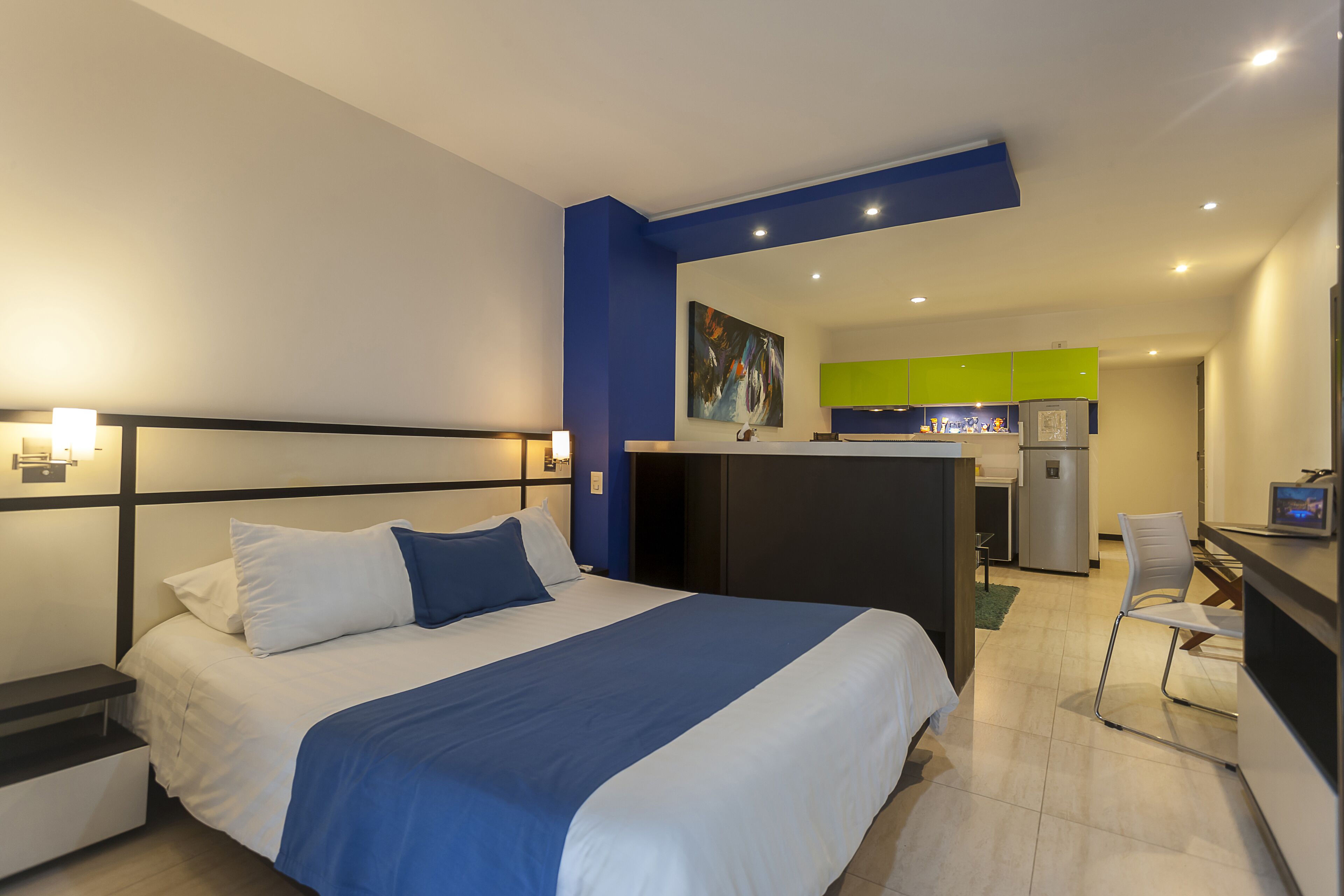 A hotel room with a bed, bedside table, and a kitchenette area with a refrigerator and sink.