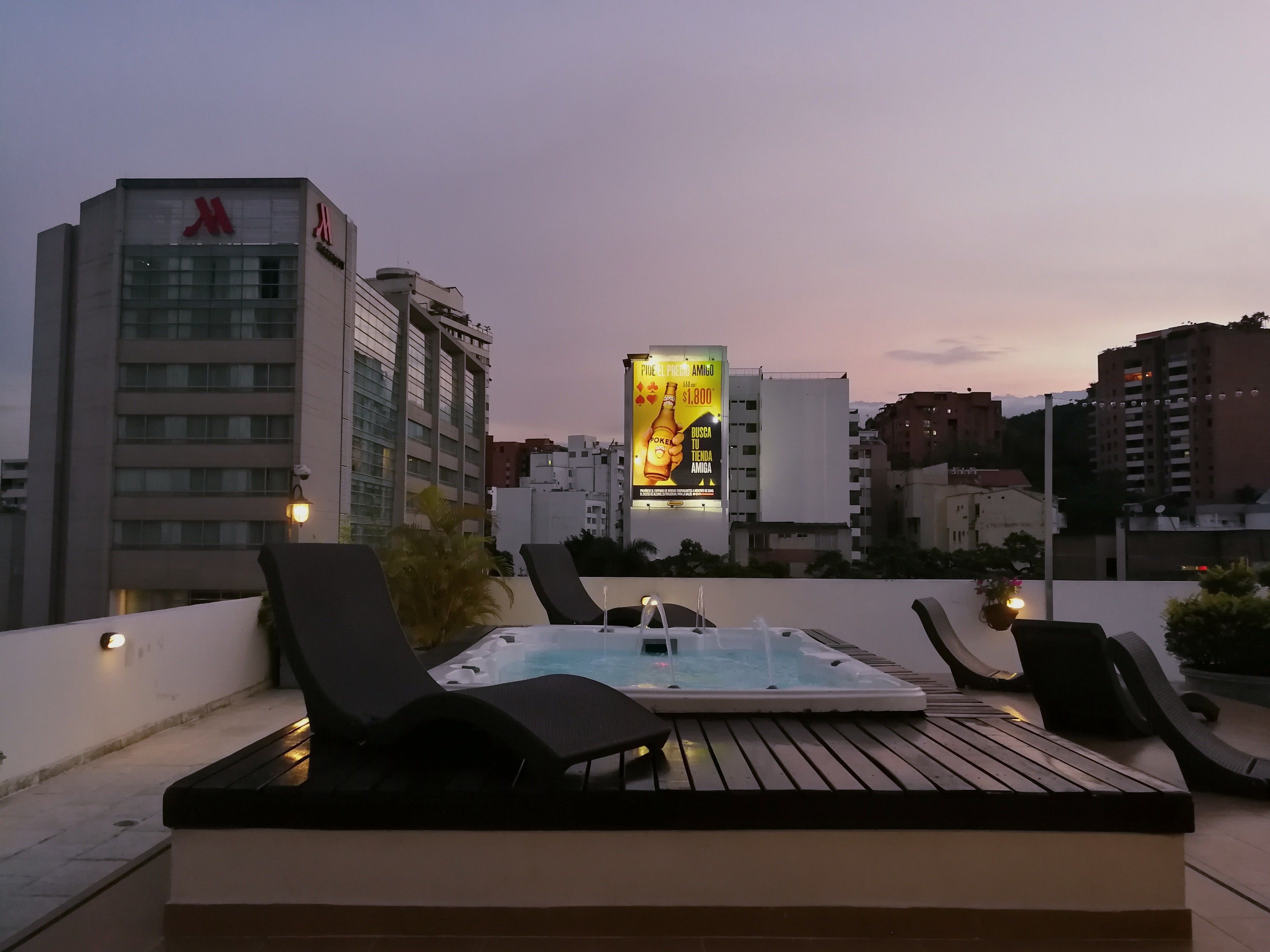 A rooftop terrace with lounge chairs, a hot tub, and a view of city buildings at dusk.