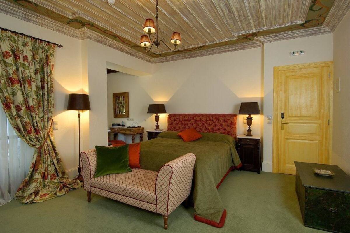A bedroom with a bed, a chair, two nightstands with lamps, a wooden door, and a green carpet.