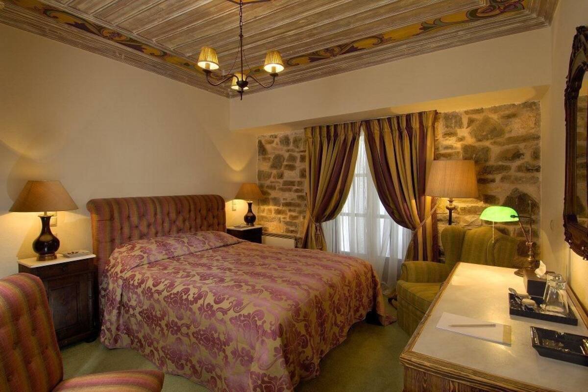 A hotel room with a large bed, two bedside tables with lamps, a green armchair, a desk with a lamp, and a stone wall.