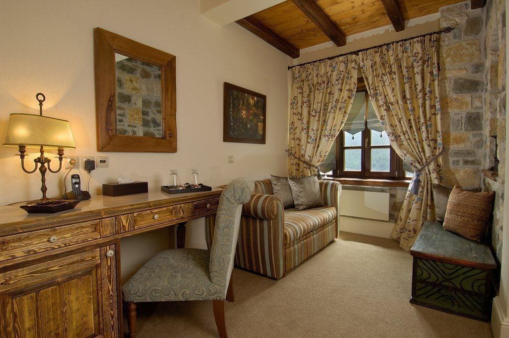 A cozy room with a wooden desk, a cushioned bench, a patterned armchair, and a stone wall.