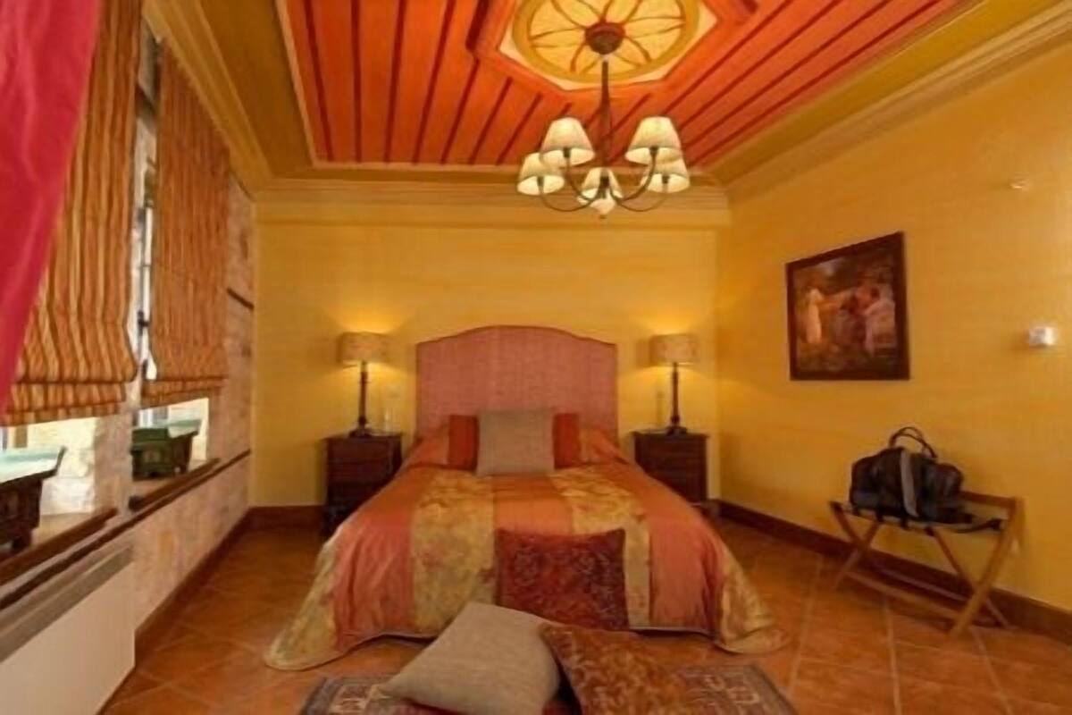 A hotel room with a large bed, two bedside lamps, a chandelier, a painting on the wall, and a suitcase on a chair.