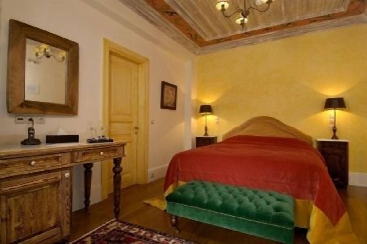 A bedroom with a wooden dresser, a mirror, a bed with a red and green upholstered headboard, and two bedside lamps.
