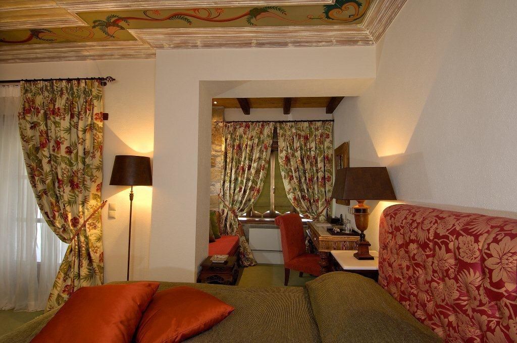 Une chambre d’hôtel avec un lit, un bureau, une chaise et un canapé. Il y a des rideaux aux fenêtres et une lampe sur le bureau.