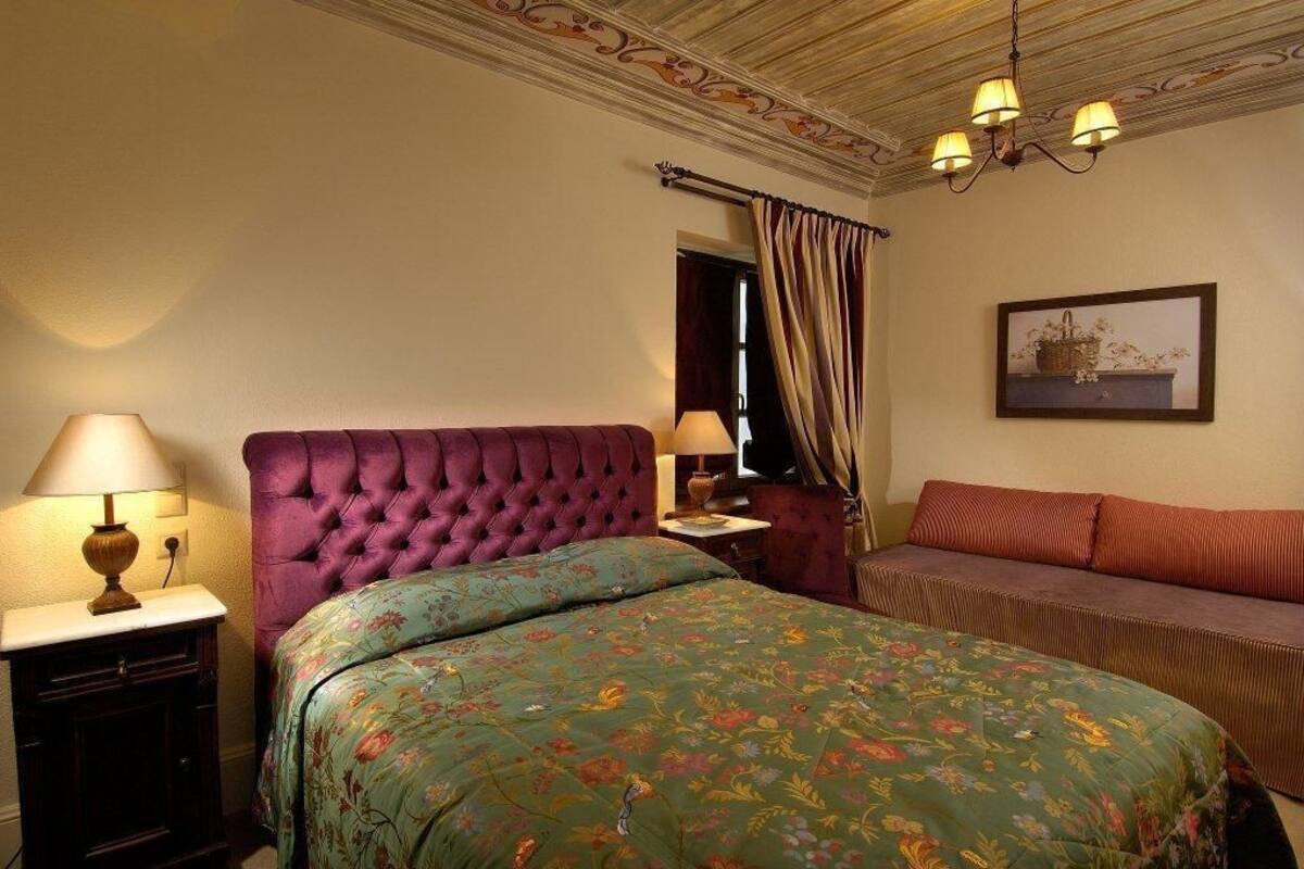 A bedroom with a tufted headboard, floral bedspread, a side table with a lamp, a sofa, and a framed picture on the wall.