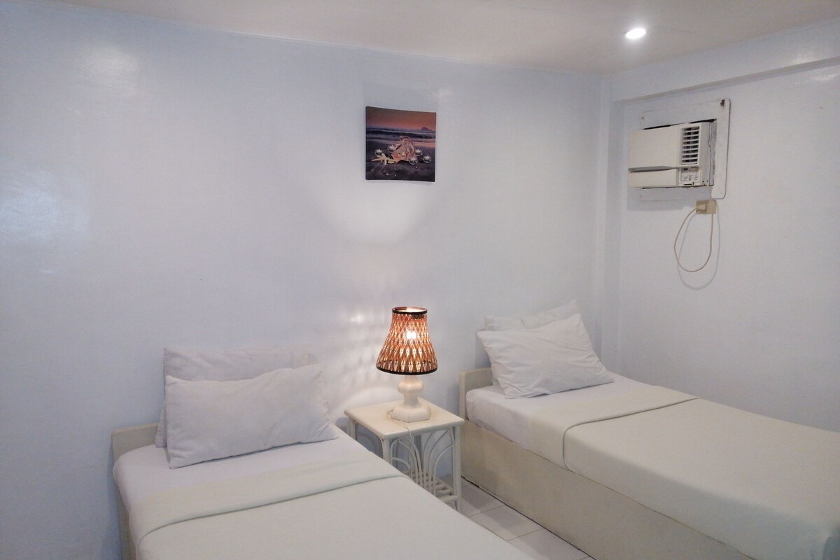 A small, clean room with two single beds, a bedside table with a lamp, a wall-mounted air conditioner, and a small picture on the wall.