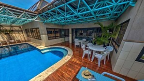 A swimming pool with a wooden deck, surrounded by white lounge chairs and tables, and a blue metal roof structure. - Hotel Massis (São Paulo)