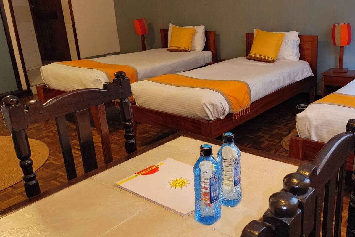 Habitación de hotel con tres camas, cada una con cabecero de madera, ropa de cama blanca y mantas naranjas. Se observa una silla de madera con estructura metálica, una mesita con un mantel individual blanco y dos botellas de agua.