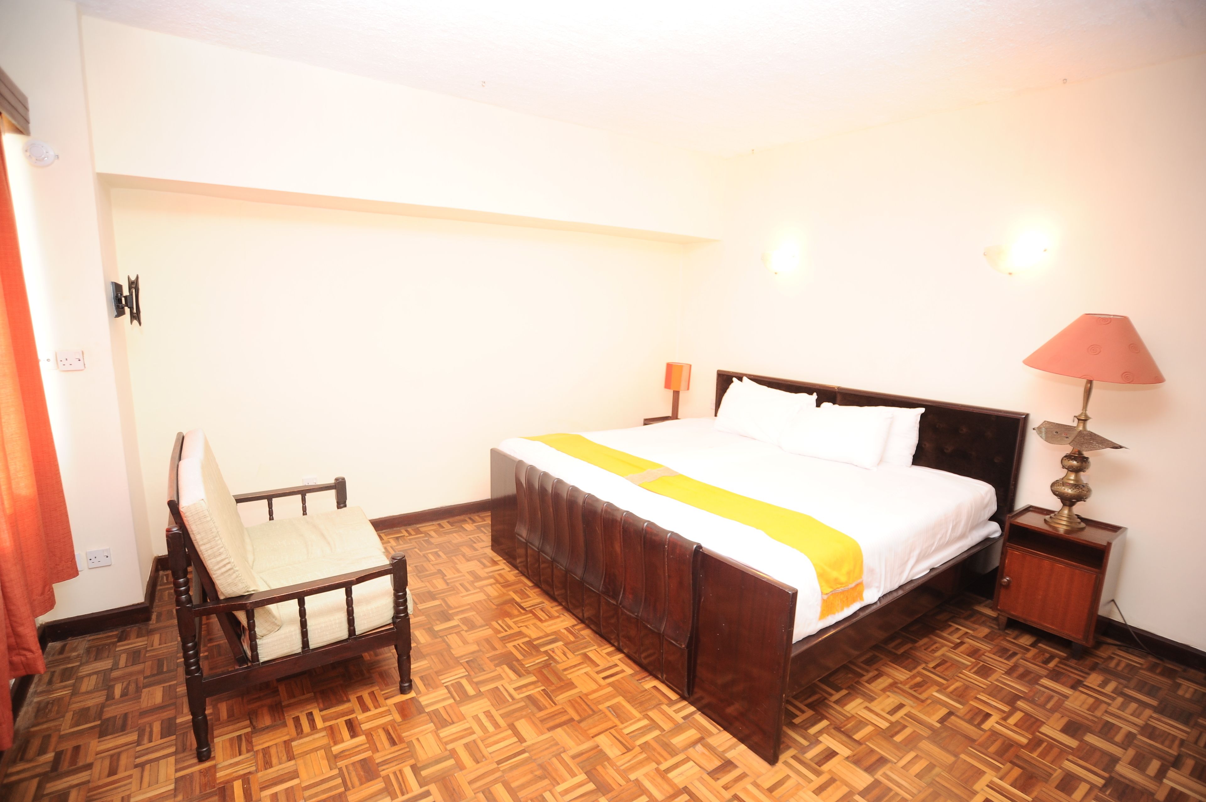 Una habitación de hotel con piso de madera, una cama con colcha blanca y amarilla, una mesita de noche de madera con una lámpara, y una silla con cojín blanco.