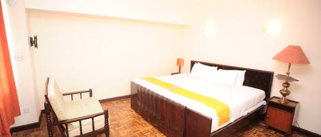 Una habitación de hotel con piso de madera, una cama con colcha blanca y amarilla, una mesita de noche de madera con una lámpara, y una silla con cojín blanco.