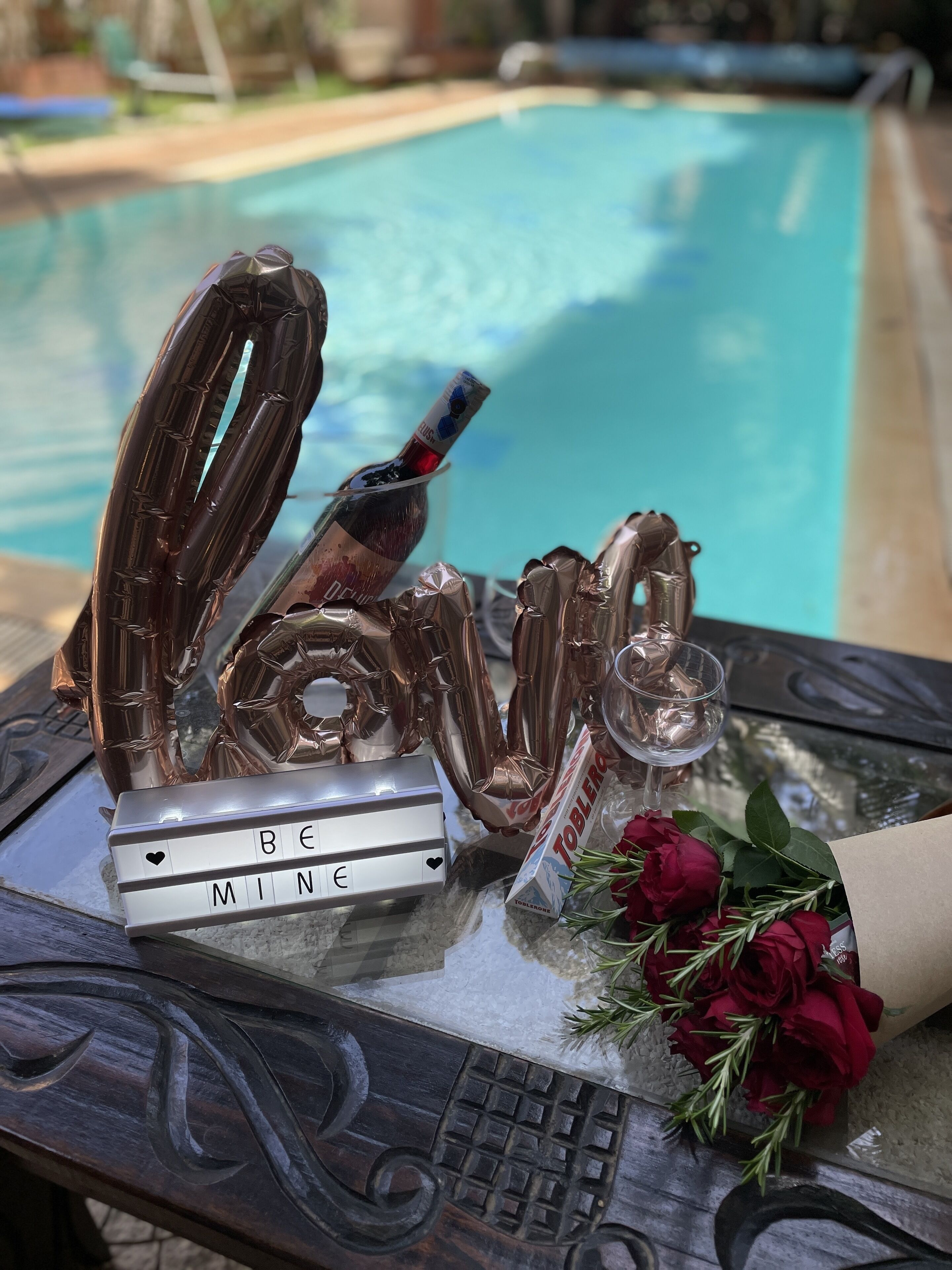 A table with a decorative arrangement including a bottle, a light box with the text "BE MINE", a glass, and a bouquet of roses.