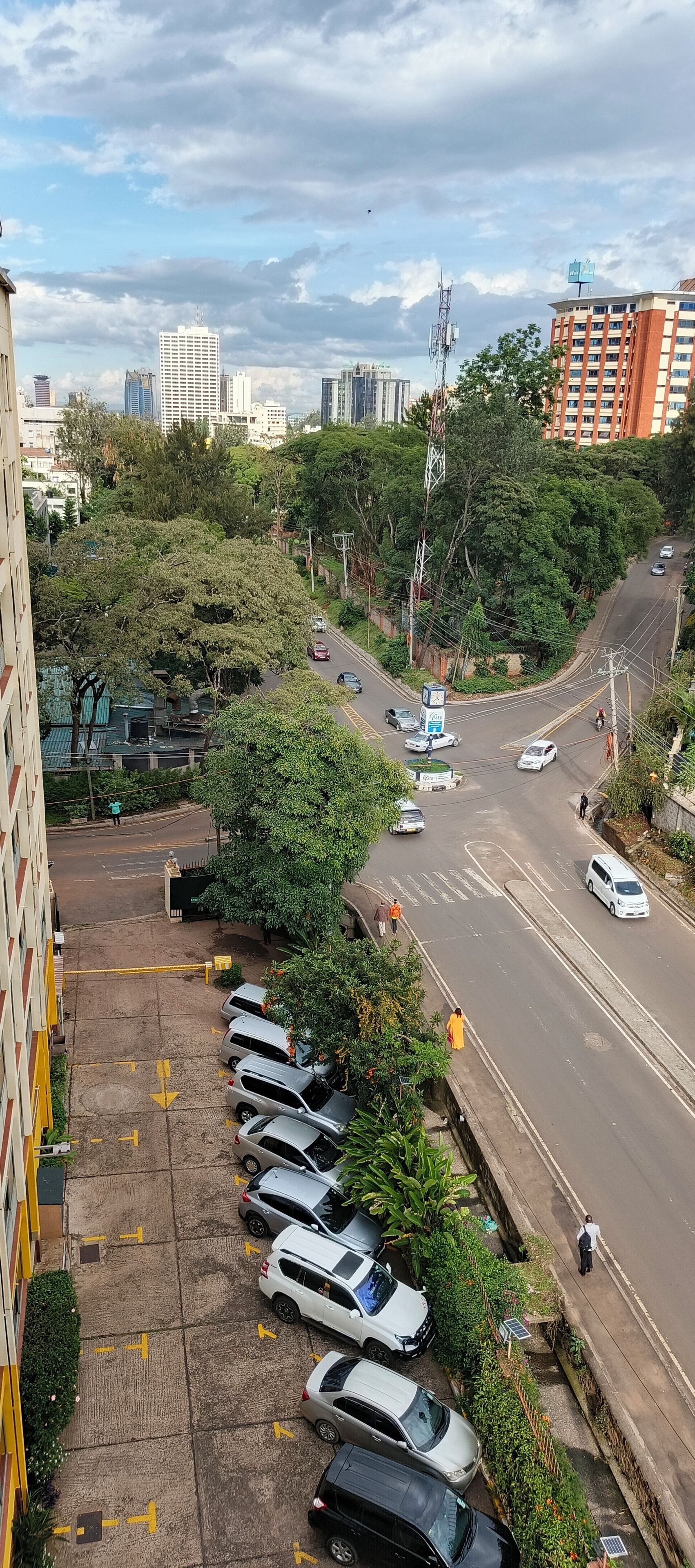 A parking area with multiple cars, a road with vehicles, and buildings in the background.