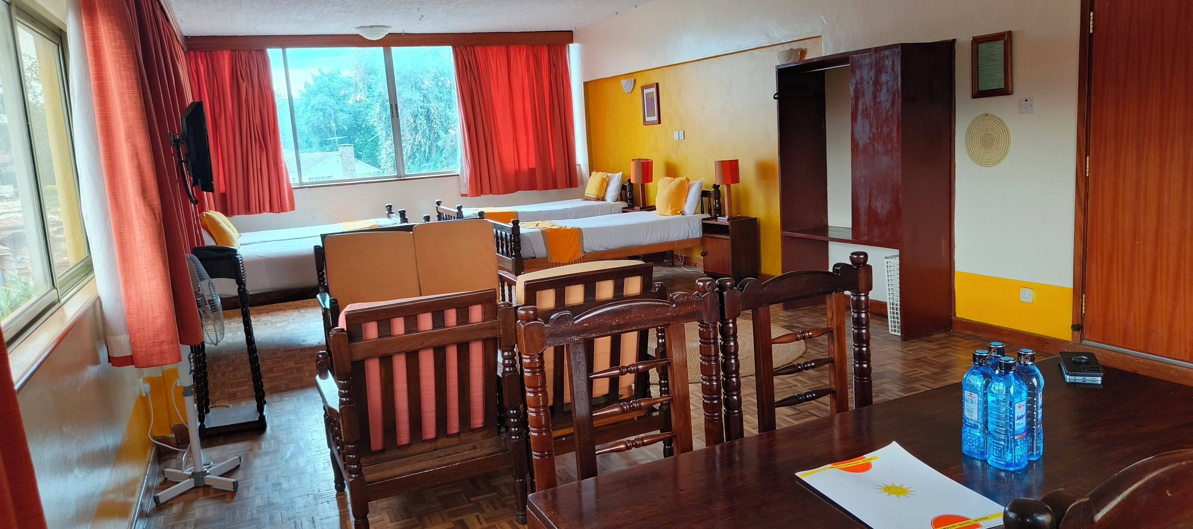 Une chambre d’hôtel avec deux lits, une table à manger, des chaises, un ventilateur et une fenêtre avec des rideaux.