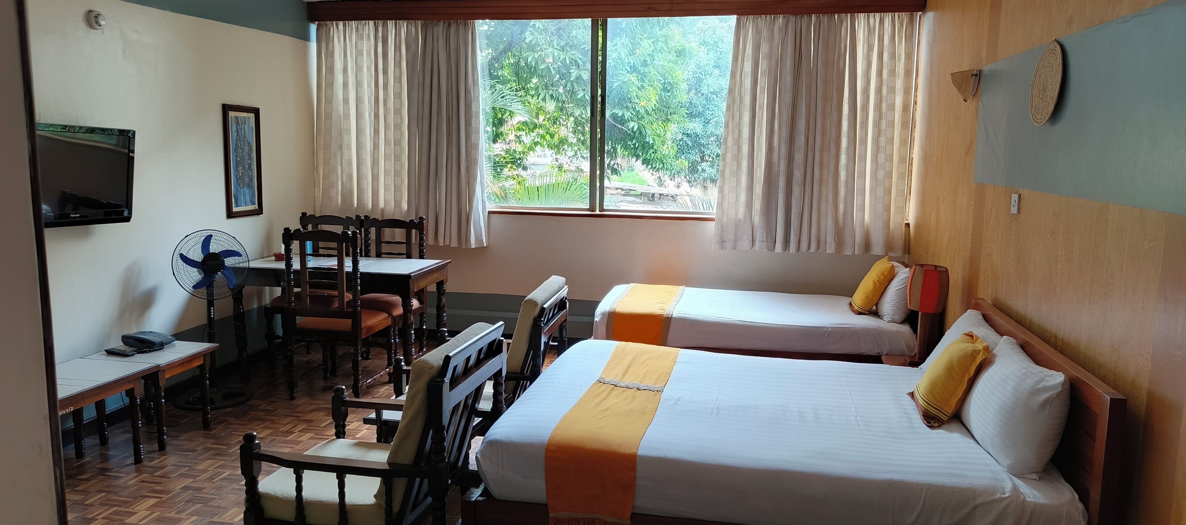 A hotel room with two beds, a desk, a chair, a fan, and a window with curtains.
