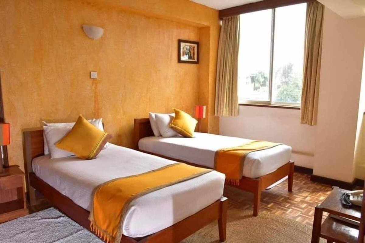 Habitación de hotel con dos camas individuales, un cabecero de madera, un amplio ventanal con cortinas y una pequeña mesa con lámpara.