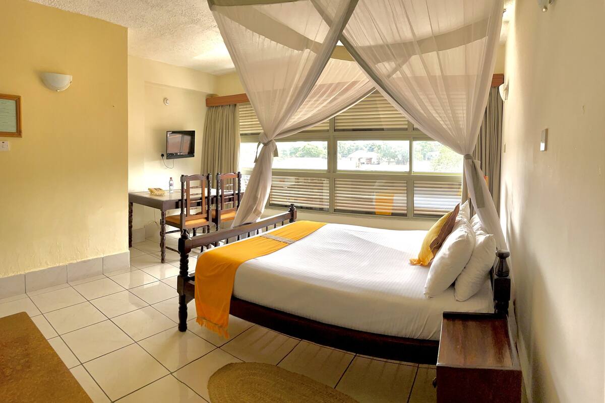 Habitación de hotel con cama, escritorio, silla, televisor y ventana con cortinas.
