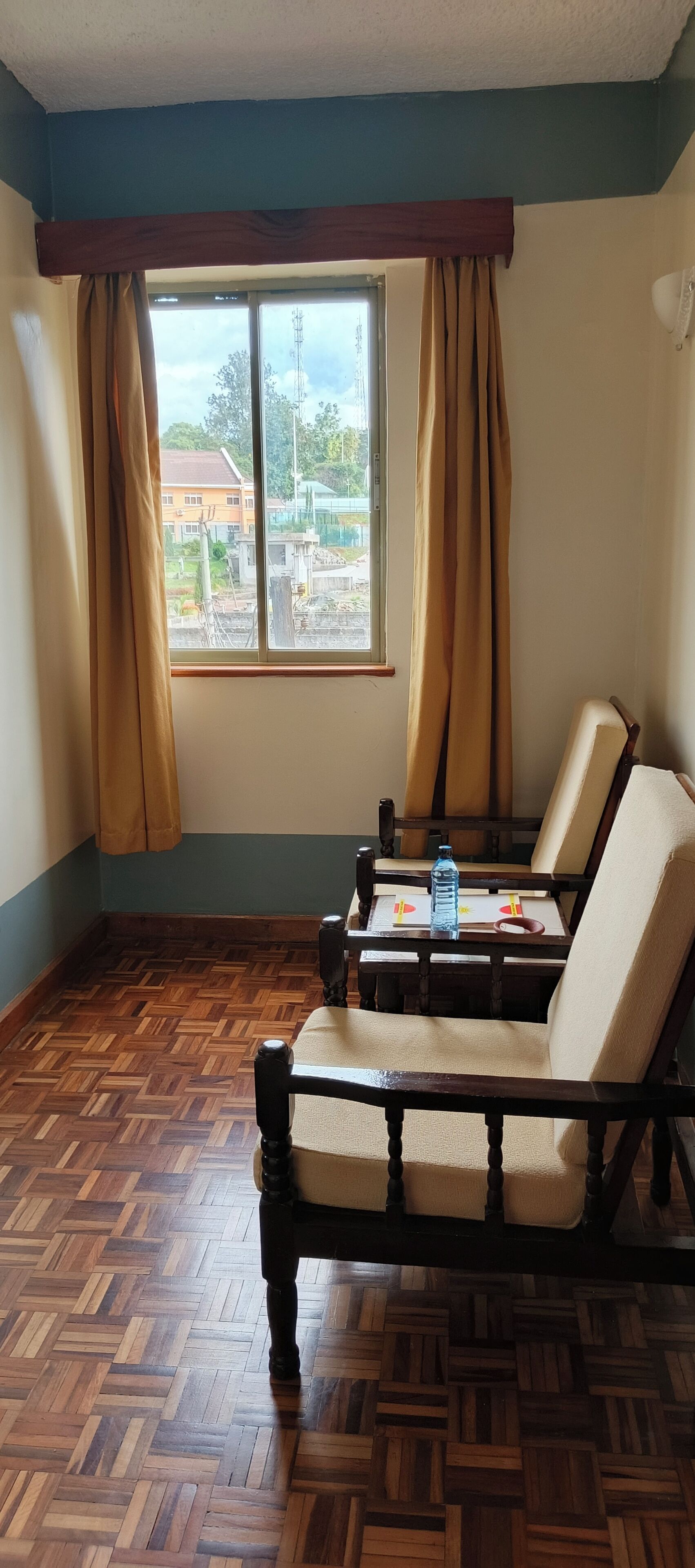 A room with a wooden herringbone floor, two wooden chairs with cushions, a window with curtains, and a view of a construction site outside.